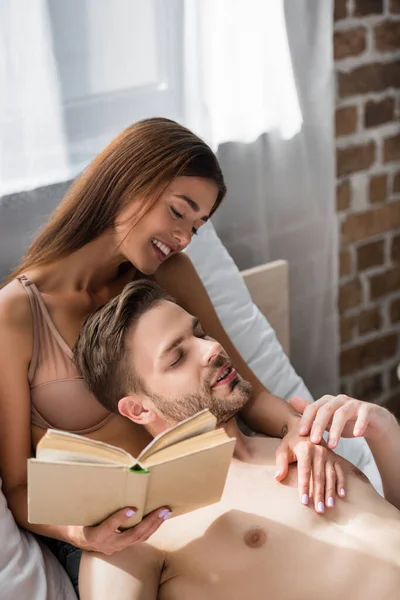 Улыбающаяся женщина, держащая книгу и трогающая парня без рубашки, лежащего на коленях — стоковое фото