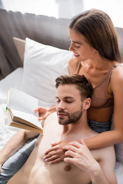 Seductive woman touching shirtless boyfriend while reading book in bedroom — Stock Photo