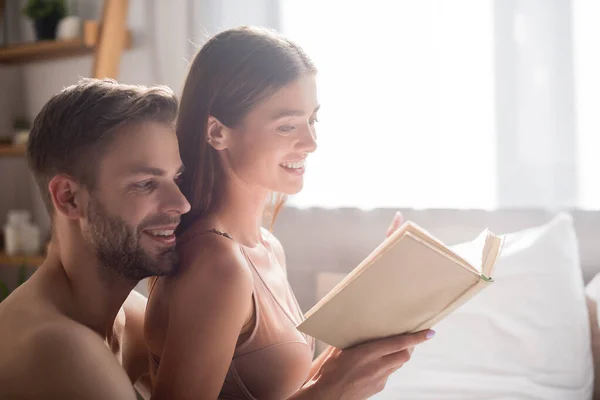 Alegre sin camisa hombre detrás seductora novia lectura libro en dormitorio - foto de stock
