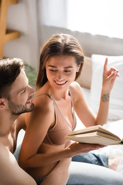 Mujer alegre en sujetador sosteniendo libro y señalando con el dedo cerca del hombre sin camisa en el dormitorio - foto de stock