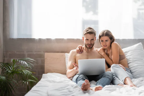 Mujer conmocionada tocando novio sin camisa usando el ordenador portátil en la cama - foto de stock