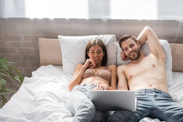 Sexy sonriente pareja acostada en la cama y el uso de ordenador portátil juntos - foto de stock