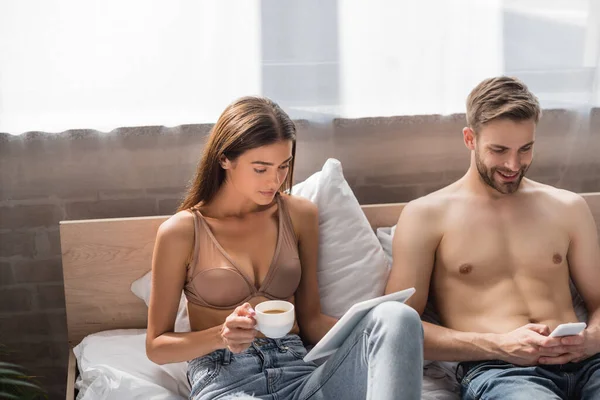 Hemdloser Mann meldet sich mit Tablet und Kaffeetasse auf Smartphone bei Freundin in BH — Stockfoto