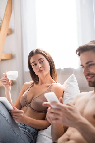 Mujer sexy sosteniendo la taza de café y tableta digital mientras mira al hombre sin camisa charlando en el portátil en primer plano borroso - foto de stock