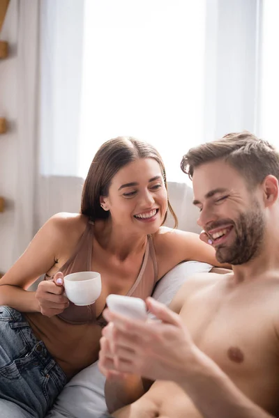Selektiver Fokus der Frau, die eine Tasse Kaffee in der Hand hält, während sie fröhliche Freund-Nachrichten auf dem Handy im verschwommenen Vordergrund betrachtet — Stockfoto