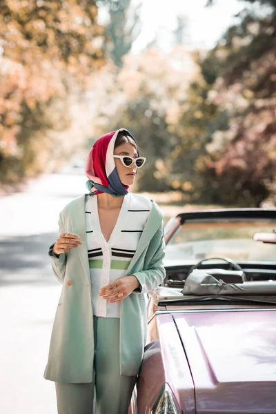 Модная женщина в платке и солнцезащитных очках стоит рядом с машиной на дороге — стоковое фото