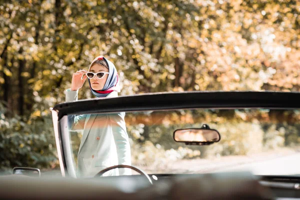 Elegante donna in occhiali da sole in piedi vicino cabriolet auto in primo piano offuscata all'aperto — Foto stock