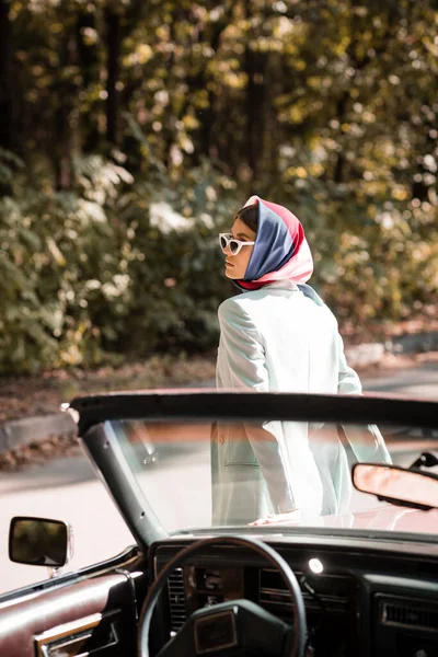 Femme en foulard et lunettes de soleil debout près de l'auto sur le premier plan flou sur la route — Photo de stock