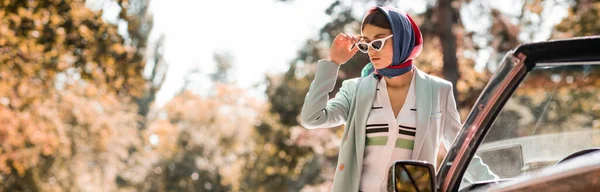 Mujer joven sosteniendo gafas de sol cerca de coche al aire libre, pancarta - foto de stock