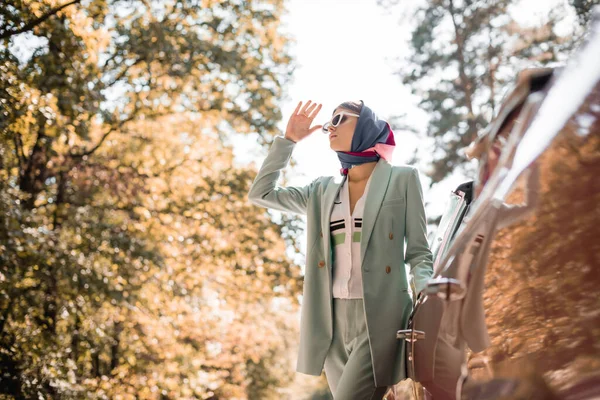 Mujer de moda en gafas de sol mirando hacia otro lado cerca del coche al aire libre en primer plano borroso - foto de stock