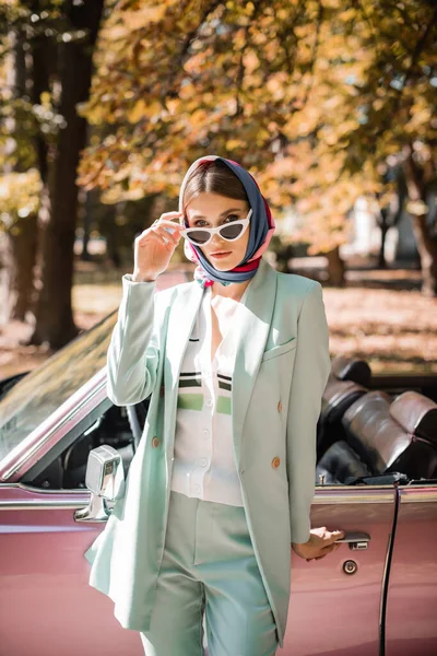 Mujer con estilo en gafas de sol mirando a la cámara cerca de coches antiguos al aire libre - foto de stock