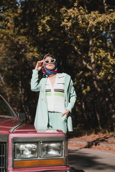 Mujer con estilo en el pañuelo tocando gafas de sol cerca de coche en primer plano borrosa en la carretera - foto de stock