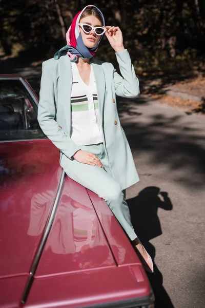 Jovem mulher em óculos de sol sentado no carro vintage com estrada no fundo — Fotografia de Stock