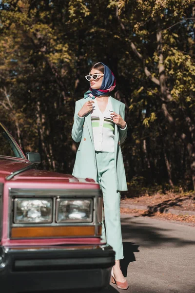 Mulher na moda de pé perto retro auto em primeiro plano desfocado na estrada — Fotografia de Stock