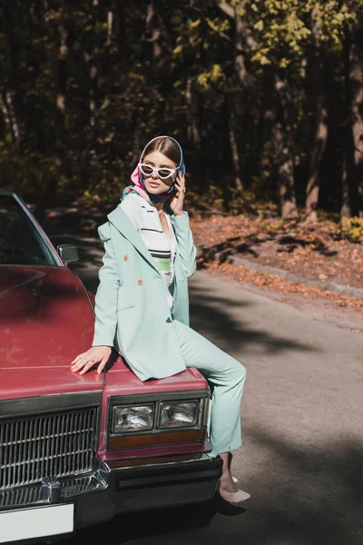 Стильна жінка в сонцезахисних окулярах і окулярах сидить на бампері автомобіля біля дороги — стокове фото