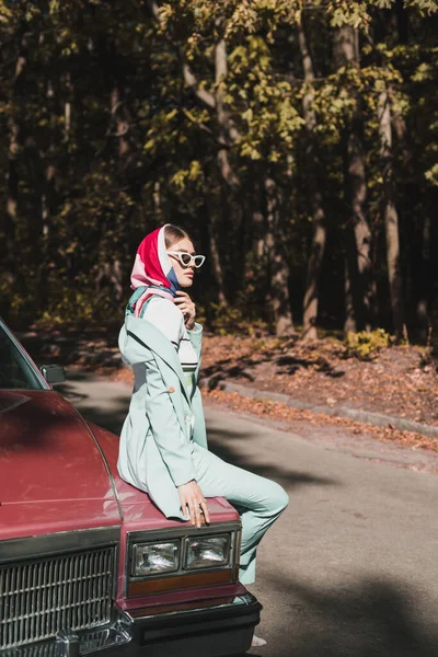 Вид сбоку модной женщины в солнцезащитных очках, сидящей на бампере автомобиля во время поездки по дороге — стоковое фото