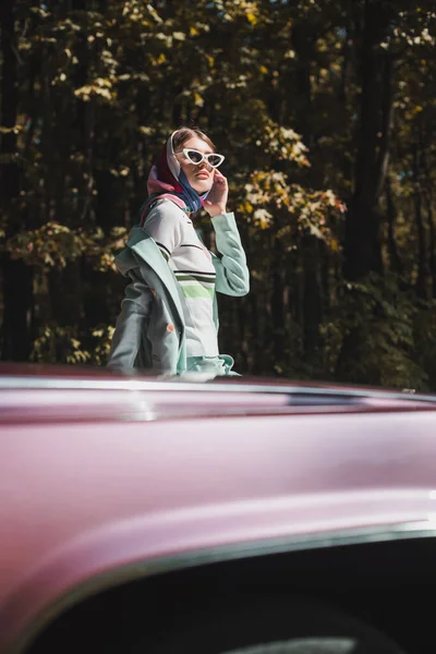 Mulher elegante em óculos de sol olhando para longe perto do carro em primeiro plano borrado ao ar livre — Fotografia de Stock
