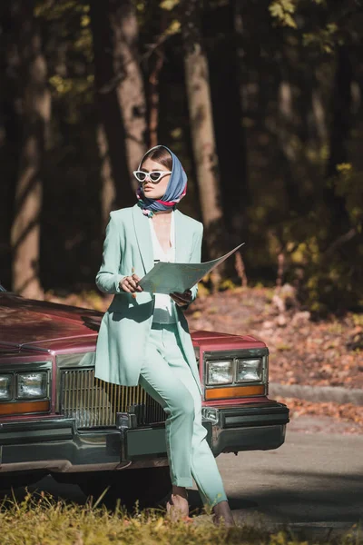 Стильна жінка в сонцезахисних окулярах тримає карту біля старовинного авто — стокове фото