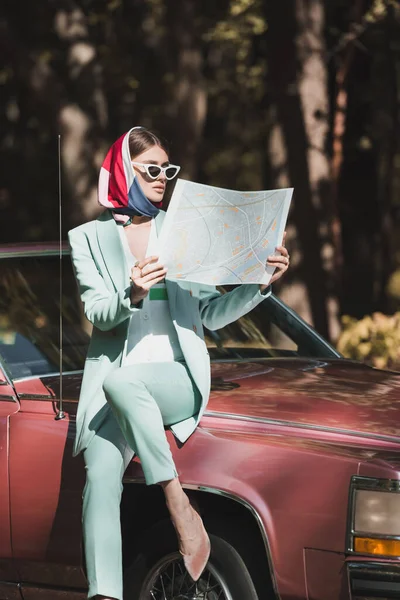 Fashionable woman in sunglasses holding map near bumper of vintage auto — Stock Photo