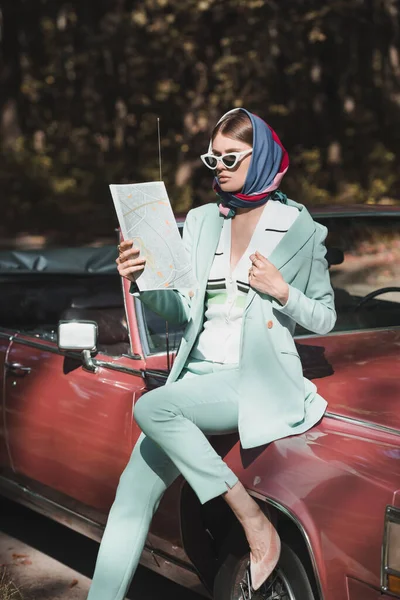 Femme élégante dans les lunettes de soleil et le foulard tenant carte tout en étant assis sur le pare-chocs de la voiture — Photo de stock