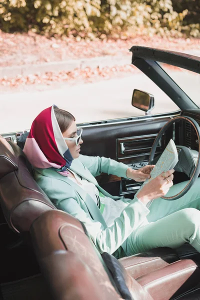 Стильна жінка тримає карту, сидячи в старовинному автомобілі на розмитому передньому плані — стокове фото
