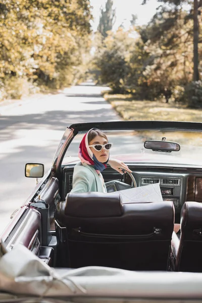 Mujer con estilo con mapa sentado en auto en primer plano borroso en la carretera durante el viaje - foto de stock