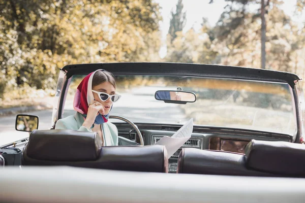 Pensive woman in sunglasses looking at map on driver seat of vintage car on blurred foreground — Stock Photo
