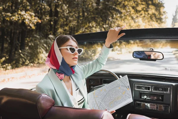 Элегантная женщина держит карту, сидя в машине без крыши на дороге на размытом переднем плане — стоковое фото