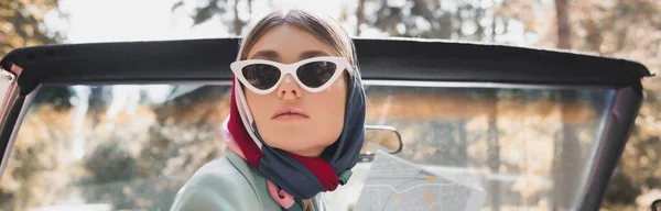 Elegante mujer en gafas de sol y pañuelo para la cabeza sosteniendo mapa en auto sin techo, pancarta - foto de stock