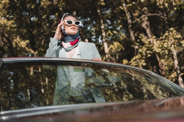 Elegant woman touching sunglasses in roofless car on blurred foreground — Stock Photo