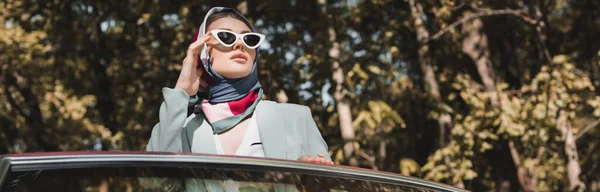 Mujer con estilo tocando gafas de sol cerca de coche al aire libre, bandera - foto de stock