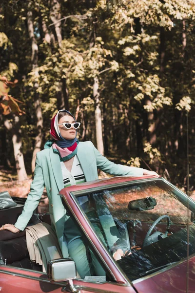 Элегантная женщина в солнечных очках смотрит вдаль от машины без крыши на размытом переднем плане — стоковое фото