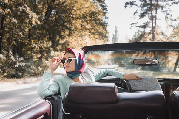 Femme élégante touchant des lunettes de soleil tout en tenant la carte en voiture rétro sur le premier plan flou — Photo de stock