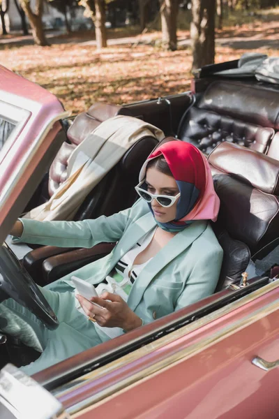 Mujer elegante en gafas de sol con smartphone mientras conduce auto vintage en primer plano borroso - foto de stock