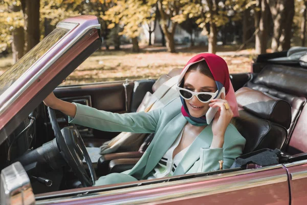 Positive woman in sunglasses talking on smartphone in roofless auto on blurred foreground — Stock Photo