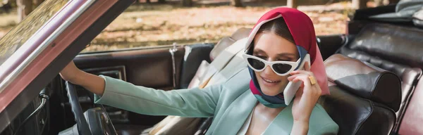 Mujer sonriente en gafas de sol hablando en el teléfono inteligente en auto vintage en primer plano borroso, bandera - foto de stock