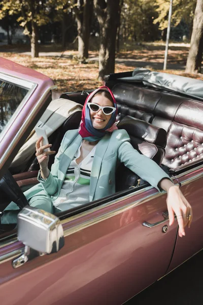 Mujer con estilo positivo que sostiene el teléfono inteligente en el asiento del conductor del coche retro en primer plano borroso - foto de stock