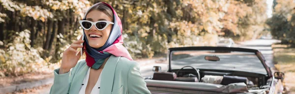 Femme souriante en lunettes de soleil parlant sur smartphone près de voiture sans toit, bannière — Photo de stock