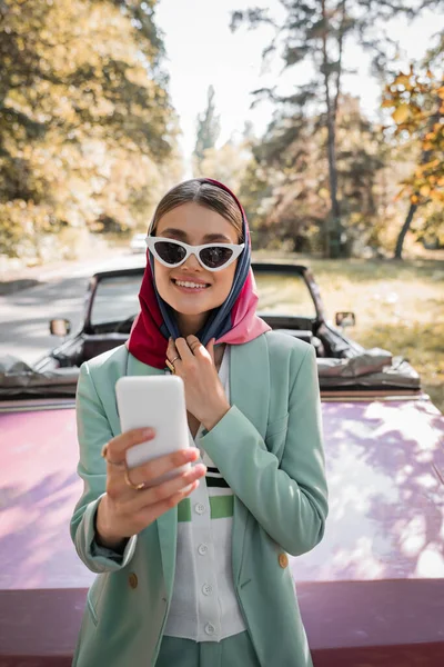 Smiling woman in sunglasses using smartphone near roofless car on blurred background — Stock Photo