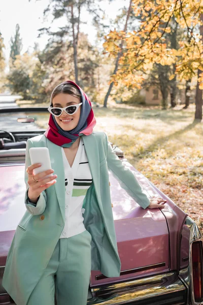 Sonriente, mujer de moda mirando el teléfono inteligente, mientras que de pie cerca de cabriolet vintage - foto de stock