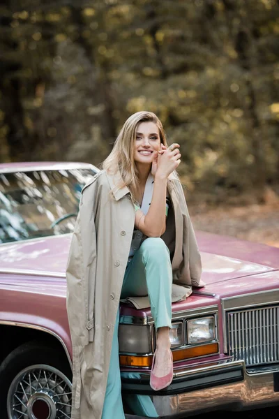 Glückliche stilvolle Frau blickt in die Kamera, während sie auf der Motorhaube eines Oldtimer-Cabriolets sitzt — Stockfoto