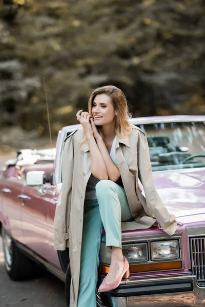 Glückliche junge Frau im Umhang, die wegschaut, während sie auf der Cabriolet-Motorhaube auf verschwommenem Hintergrund sitzt — Stockfoto