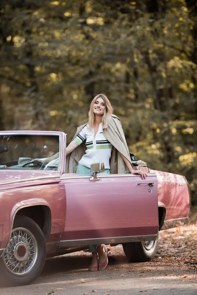 Aufgeregte Frau blickt in Kamera, während sie in der Nähe der offenen Tür eines Cabriolets steht — Stockfoto