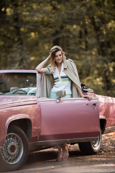Glückliche Frau im Mantel, die neben einem Retro-Cabriolet steht und die Haare berührt — Stockfoto