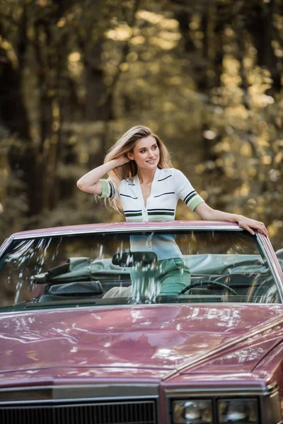 Femme heureuse regardant loin et touchant les cheveux tout en se tenant debout dans le cabriolet vintage — Photo de stock