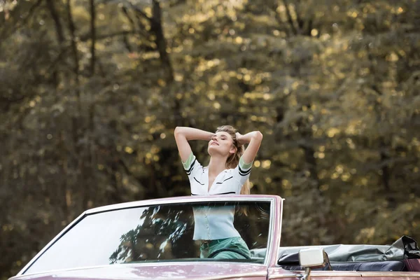 Jeune femme debout dans le cabriolet avec les yeux fermés et tenant les mains derrière la tête — Photo de stock
