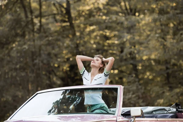 Joven mujer de pie en cabriolet con los ojos cerrados y cogidas de la mano detrás de la cabeza - foto de stock