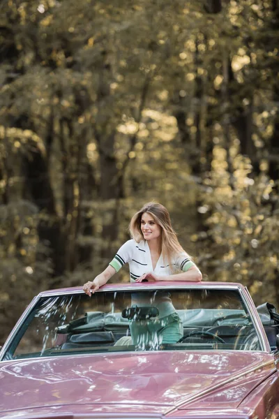 Mujer feliz mirando hacia otro lado mientras se apoya en el parabrisas del coche descapotable - foto de stock