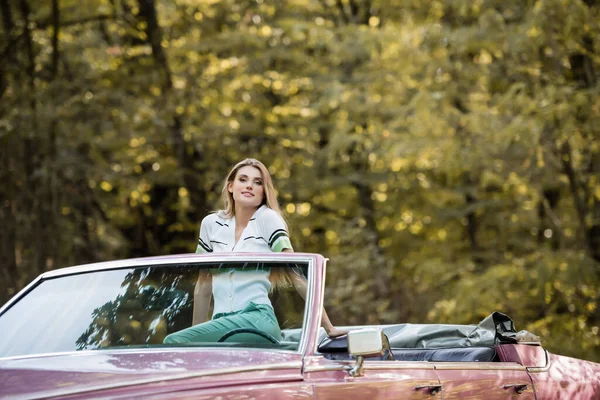 Jeune femme élégante regardant la caméra tout en posant dans la voiture décapotable vintage — Photo de stock