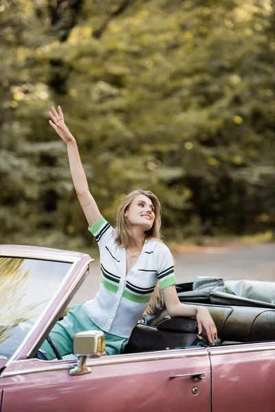 Lächelnde Frau schaut weg, während sie im Cabrio sitzt und die Hand in der Luft hält — Stockfoto